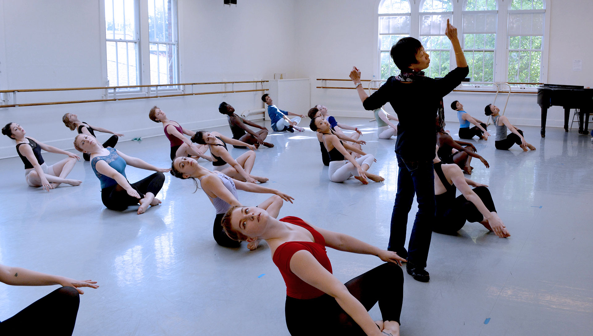 Dance students move in unison in sitting position while being directed by an instructor