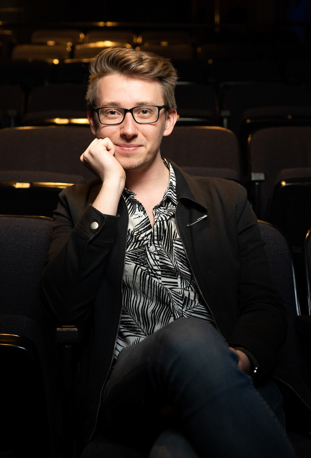 Headshot of Gavin Yehle, Cornish College of the Arts Staff. Photo of Gavin in a dark jacket and multi-colored button-up sitting in a theater seat.