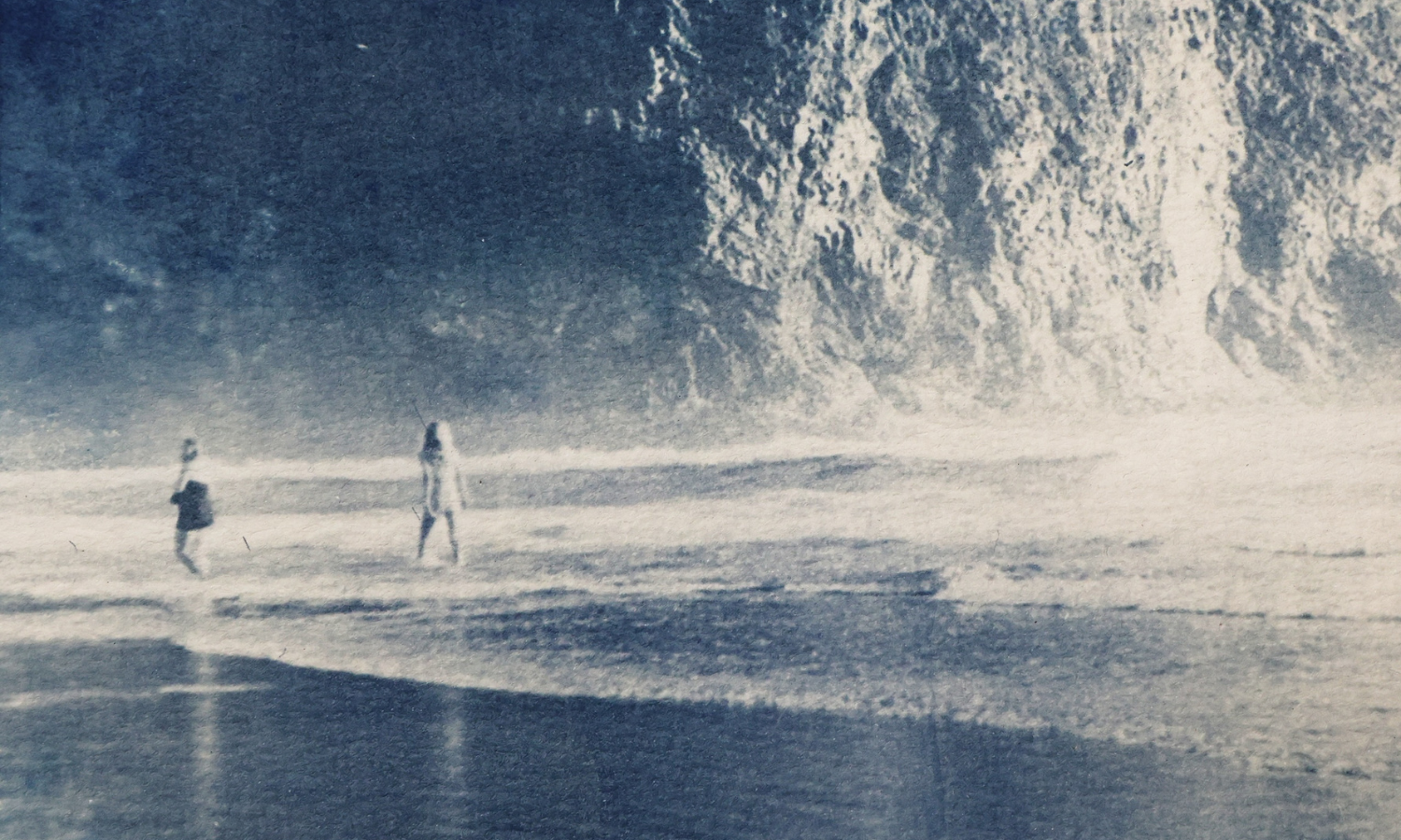 Image by Gemma Cannon-Green Blue and tan image of two people walking on a beach with a cliff in the background