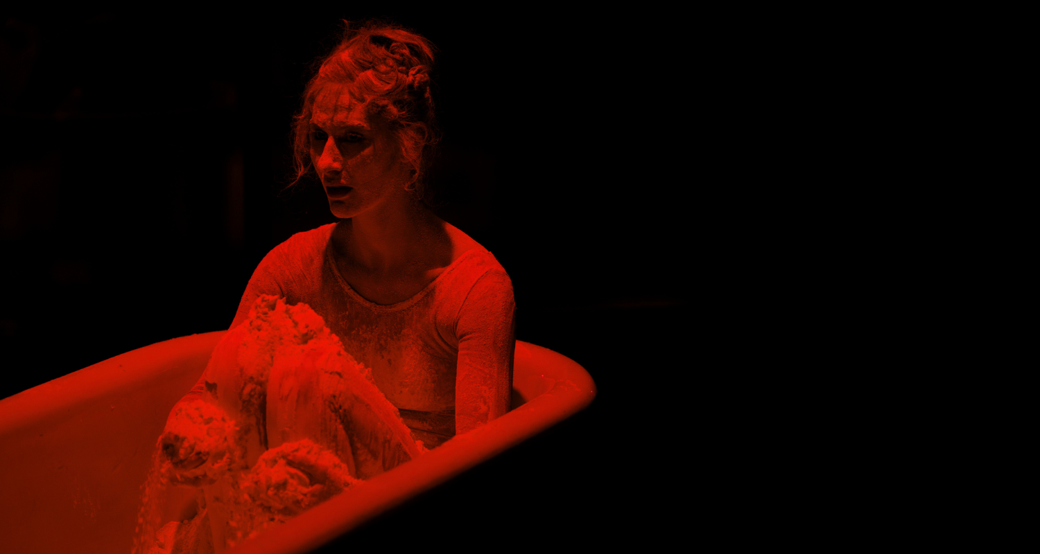 Red and black duotone photo of a woman sitting in an empty tub covered in a dusty material against a black background.