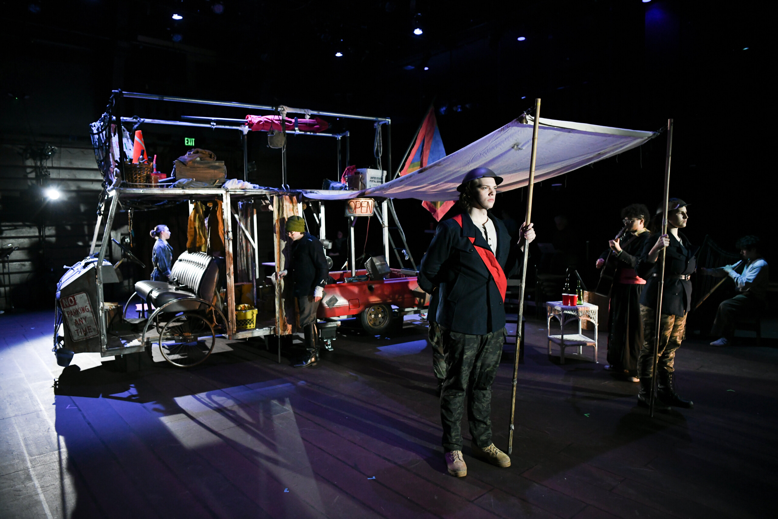 Scene from Mother Courage and Her Children; actors standing in front of a large set piece.