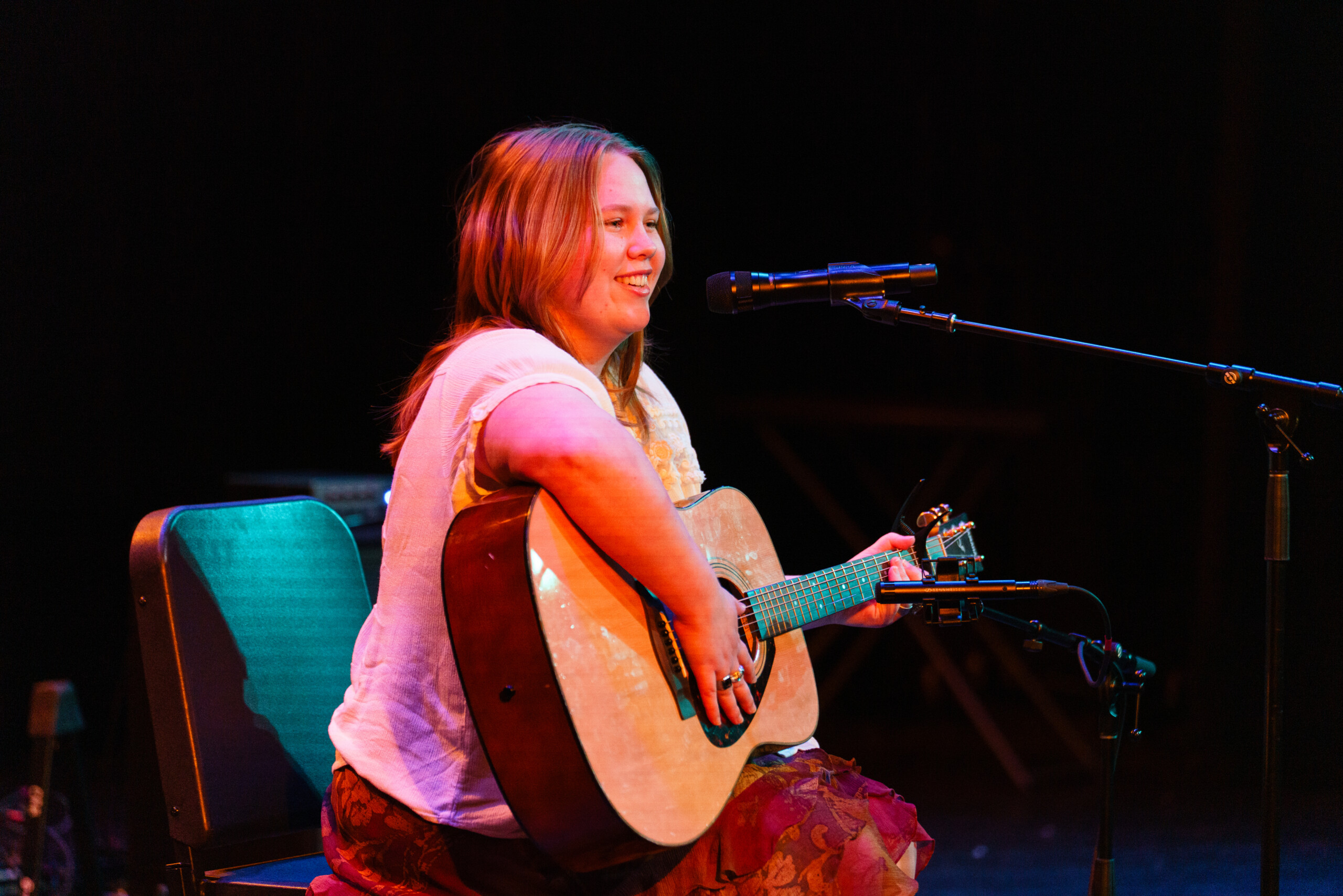 Student holding a guitar on stage while sitting.